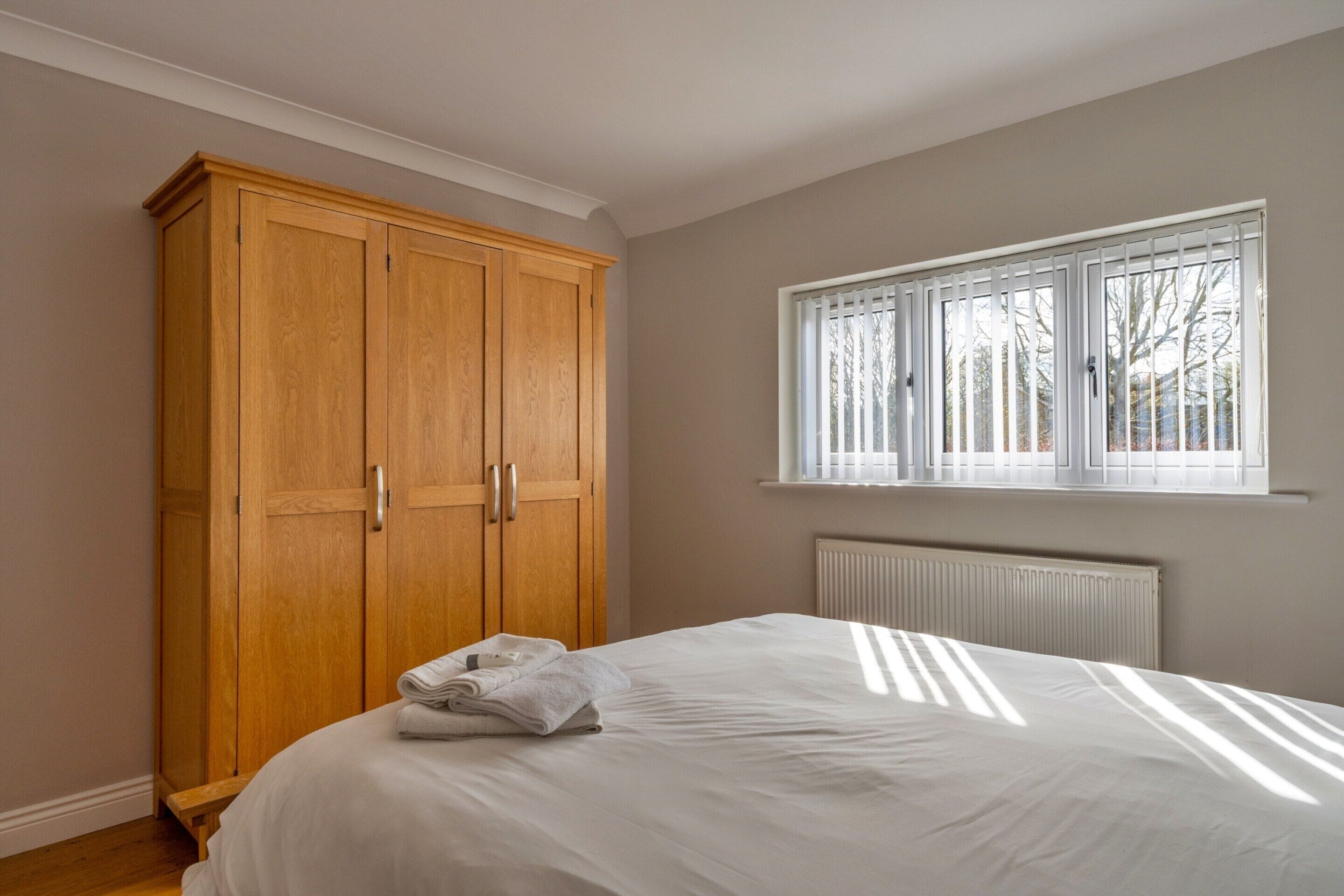 Un dormitorio con un amplio armario de madera, una cama con sábanas blancas y una ventana con persianas.