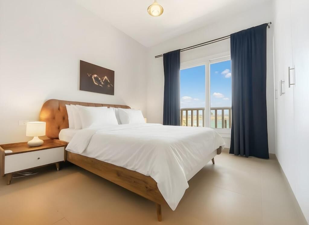 Un dormitorio con una cama de madera, ropa de cama blanca, una mesita de noche con lámpara, una obra de arte en la pared y un ventanal con cortinas.