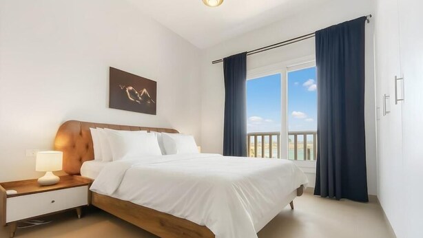 A bedroom with a wooden bed, white bedding, a nightstand with a lamp, a wall-mounted artwork, and a large window with curtains. - El Gouna luxury apartment lagoon and pool view (Hurghada 2)