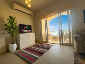 A living room with a flat-screen TV, a potted plant, a striped rug, and a sliding glass door leading to a balcony.