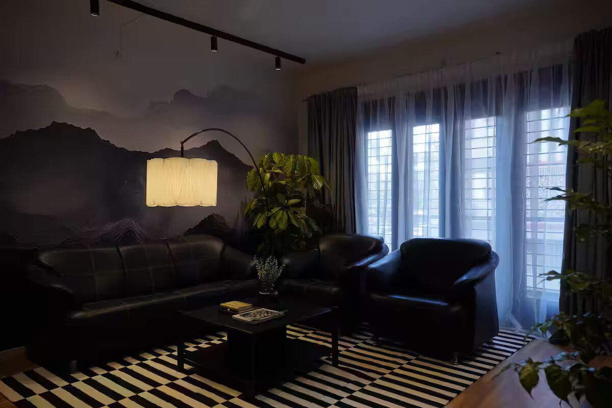 A room with a black and white checkered floor, a black sofa, a black armchair, a table with a lamp, and a large plant.