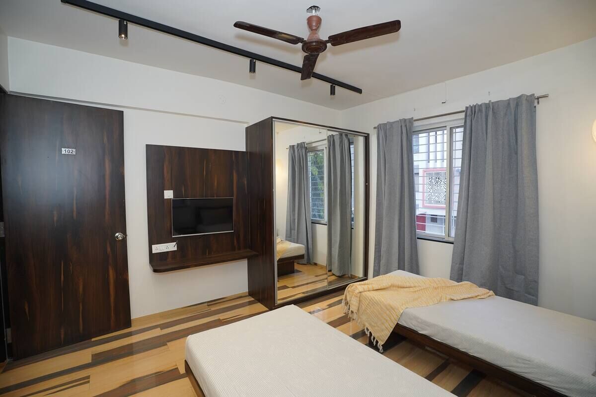 A room with a bed, a TV mounted on the wall, a ceiling fan, and a window with curtains.