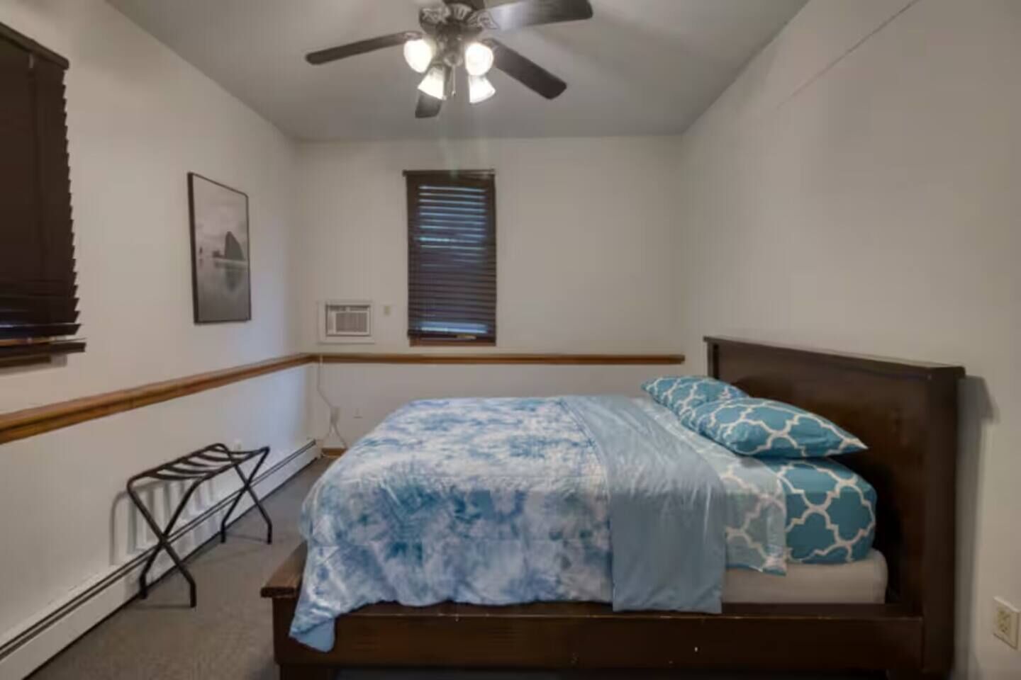 A bedroom with a bed, a ceiling fan, a window with blinds, a wall-mounted air conditioner, and a small metal rack.