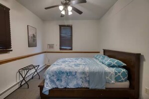 Un dormitorio con cama, ventilador de techo, ventana con persianas, aire acondicionado de pared y una pequeña estantería metálica.