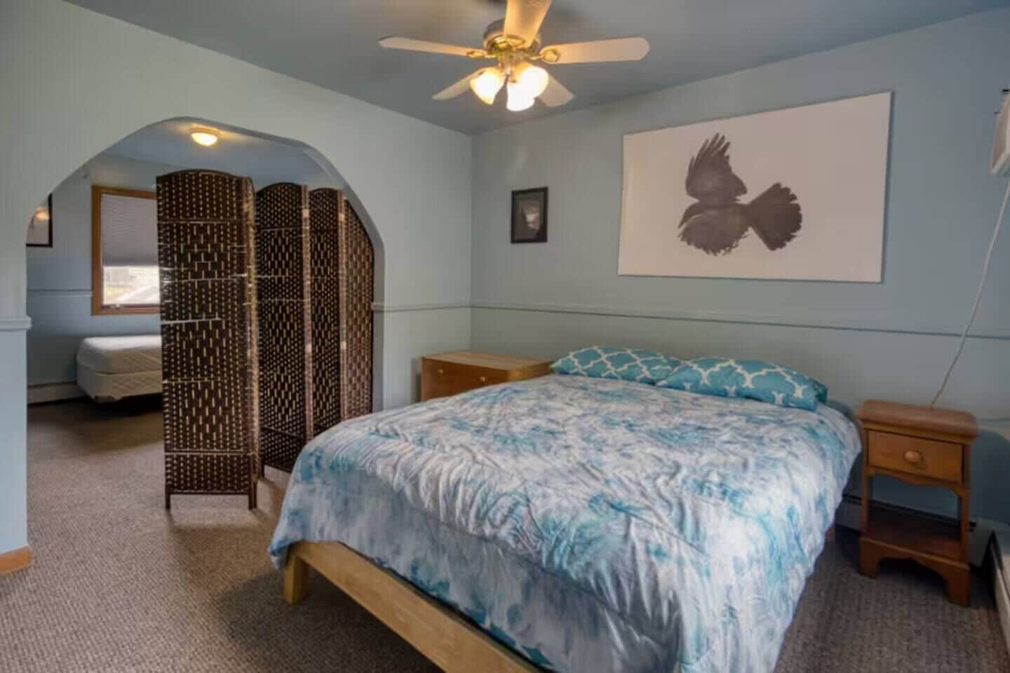 A bedroom with a bed, a nightstand, a ceiling fan, a wall-mounted artwork, and a window with blinds.