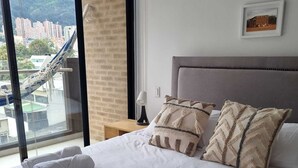 A neatly made bed with white linens and decorative pillows, a bedside table with a lamp, and a large window offering a view of a cityscape and mountains. - Cozy and sophisticated apartment with a pool, gym, and spa in the Financial District (Bogotá)