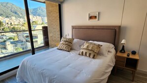 A hotel room with a large bed, a nightstand, a lamp, and a view of a cityscape through a large window. - Cozy and sophisticated apartment with a pool, gym, and spa in the Financial District (Bogotá)