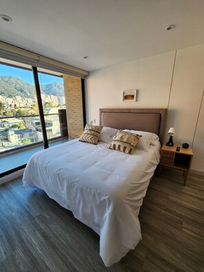 A hotel room with a large bed, a nightstand, a lamp, and a view of a cityscape through a large window. - Cozy and sophisticated apartment with a pool, gym, and spa in the Financial District (Bogotá)