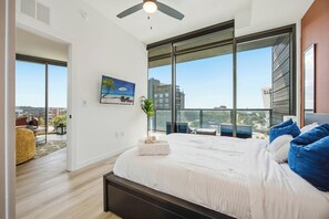 A modern bedroom with a large bed, a ceiling fan, and a view of the city through floor-to-ceiling windows.
