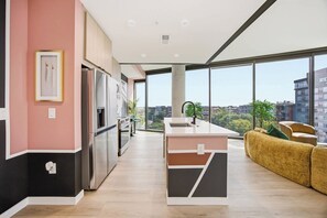 A modern kitchen with a built-in oven, a refrigerator, and a sink. There is a dining area with a sofa and a view of the city through large windows.