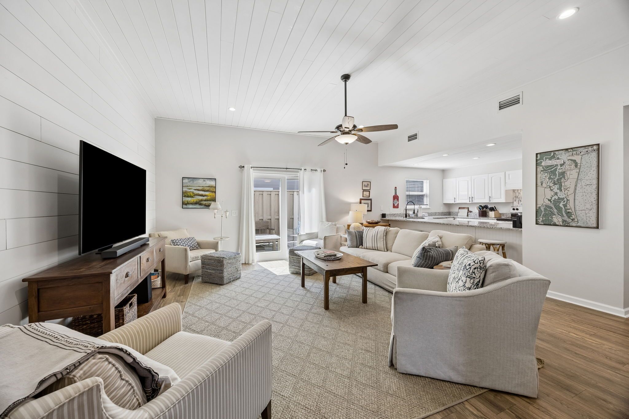 A spacious living room with a flat-screen TV, a ceiling fan, and a dining area with a kitchen in the background.