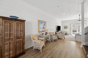 A spacious living room with a wooden floor, white walls, and a large wooden cabinet. There are wicker chairs and a small table with a lamp. A painting of a beach scene hangs on the wall. - 3BR/3BA Multilevel Condo | Steps to Pool & Beach | Monthly Stays (Fernandina Beach)
