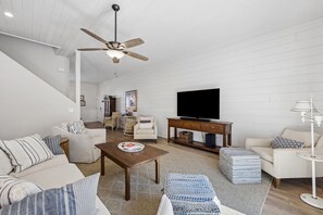 A spacious living room with a high ceiling fan, a flat-screen TV, and a dining area with wicker furniture. - 3BR/3BA Multilevel Condo | Steps to Pool & Beach | Monthly Stays (Fernandina Beach)