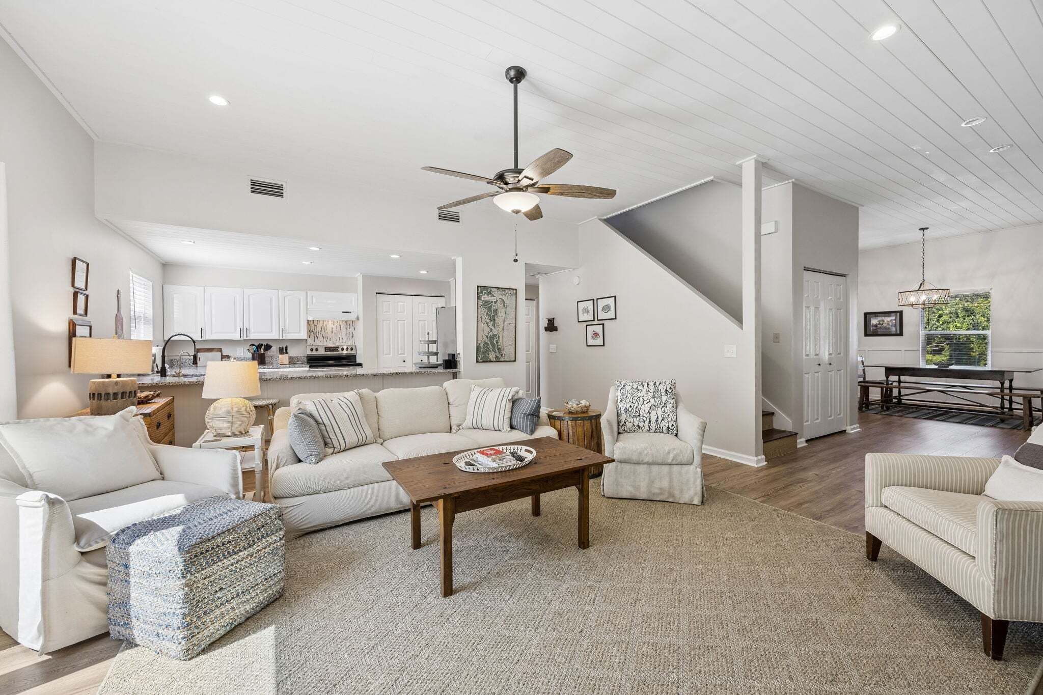 A spacious living room with a high ceiling, a ceiling fan, a dining area with a bench, and a kitchen in the background.