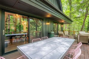 Ein Essbereich im Freien mit Tisch und Stühlen, ein Grill und Blick auf den umliegenden Wald.