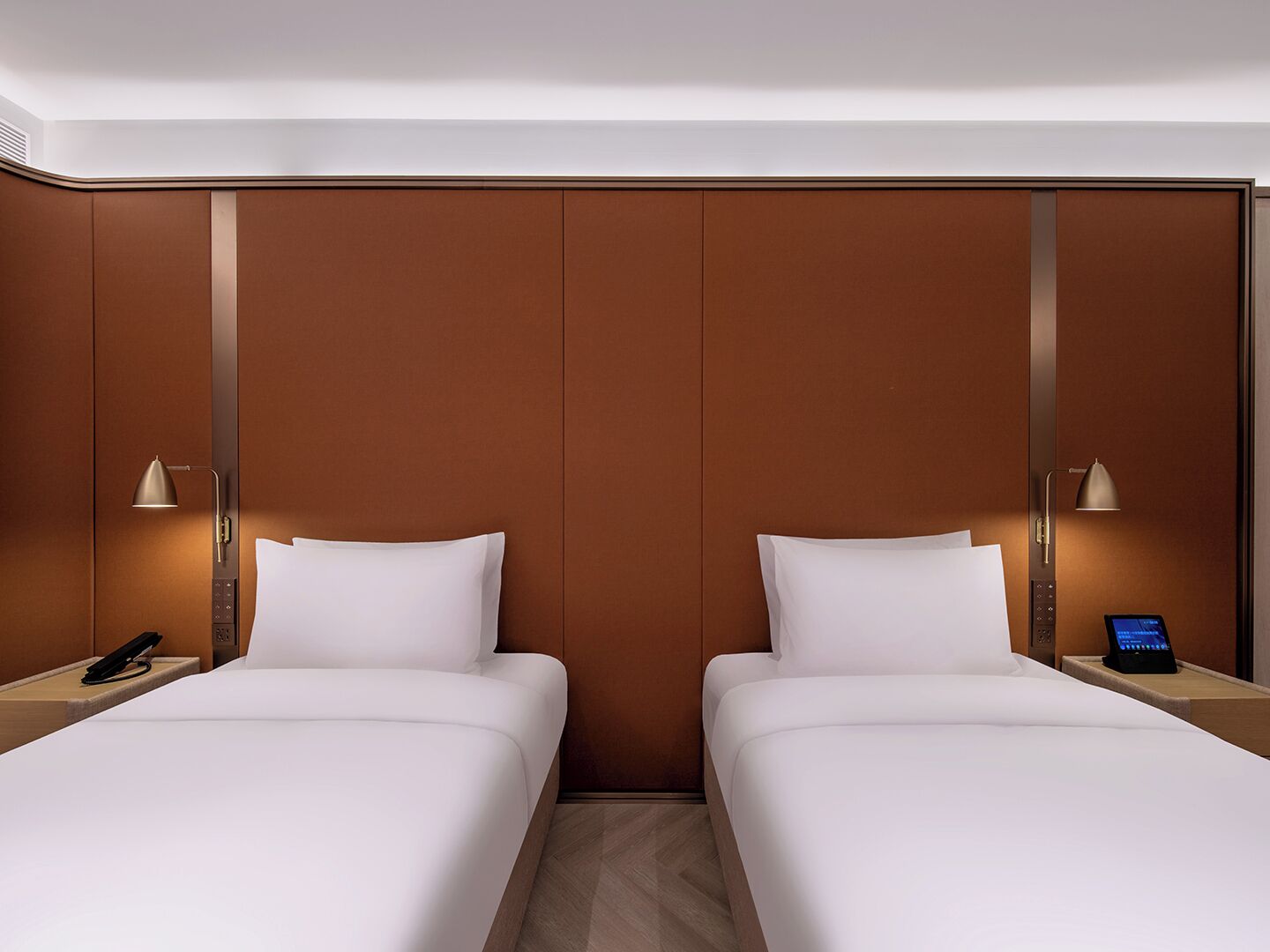 A hotel room with two beds, a bedside phone, a digital clock, and a wall-mounted lamp.