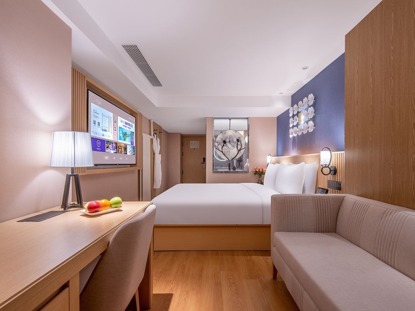 A hotel room with a bed, a desk with a laptop, a chair, a sofa, a wall-mounted TV, and a decorative mirror.