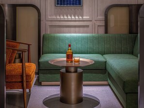A room with a green upholstered sofa, a round metal table with a bottle and glass, and a wooden chair.