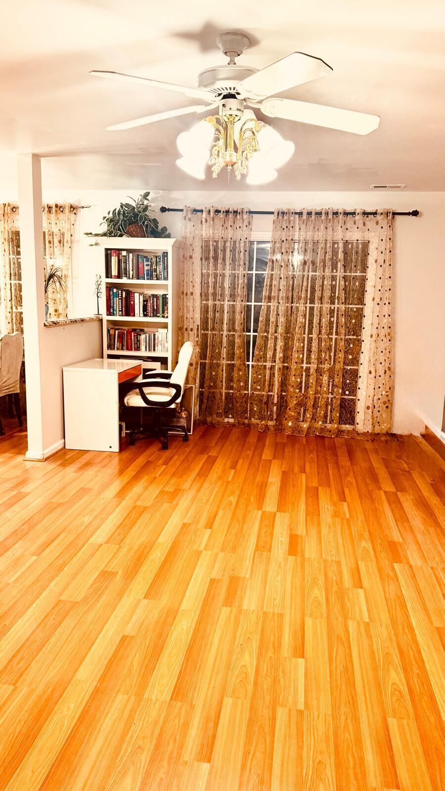 Une pièce avec un plancher en bois, un ventilateur de plafond, une bibliothèque, un bureau avec une chaise et une fenêtre avec des rideaux.