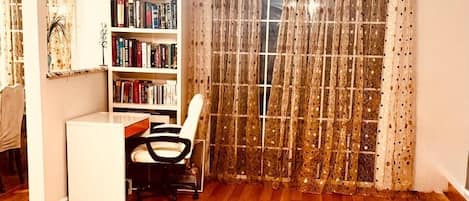 A room with a wooden floor, a ceiling fan, a bookshelf, a desk with a chair, and a window with curtains.