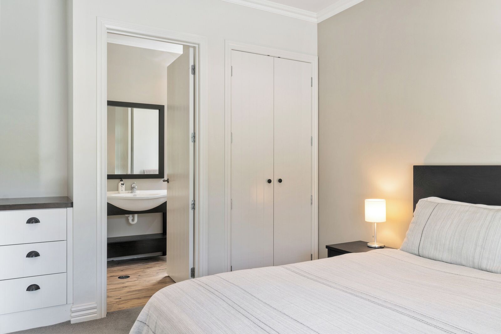 A bedroom with a bed, a nightstand, a bedside lamp, a wardrobe, and a bathroom with a sink and mirror.
