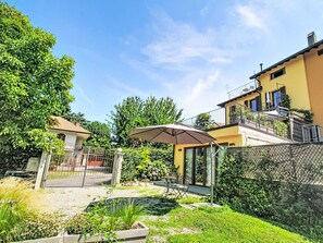 Een woonhuis met een omheinde tuin, een overdekt terras met tuinmeubilair en een geel gebouw met een balkon.