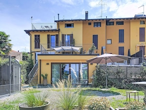 Ein zweistöckiges Gebäude mit Balkon, einer Terrasse mit Tisch und Stühlen sowie einem Garten mit verschiedenen Pflanzen.