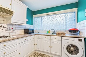 A compact kitchen with white cabinetry, a built-in microwave, a washing machine, and a window with a floral curtain.