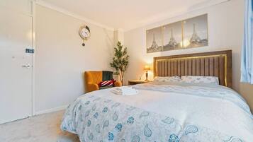 Hotellihuone, jossa on sänky, yöpöytä, lamppu, kello ja seinälle ripustettu taideteos.