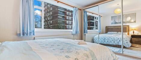A bedroom with a large bed, a mirror, a window with curtains, and a view of buildings outside.