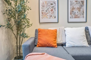 Ein graues Sofa mit drei Kissen, einer Topfpflanze und zwei gerahmten Kunstwerken an der Wand.