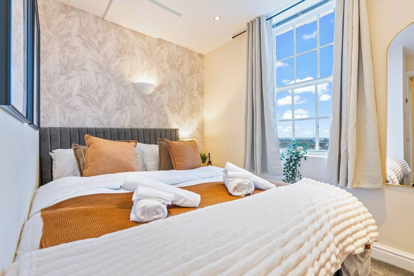 A hotel room with a large bed, a headboard, a wall-mounted light, a window with curtains, and a view of the sky.