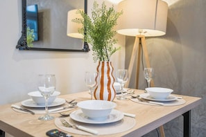 Ein Esstisch ist gedeckt mit weißen Tellern, Schüsseln und Besteck, dazu eine Vase mit Grünpflanzen und im Hintergrund zwei Lampen.