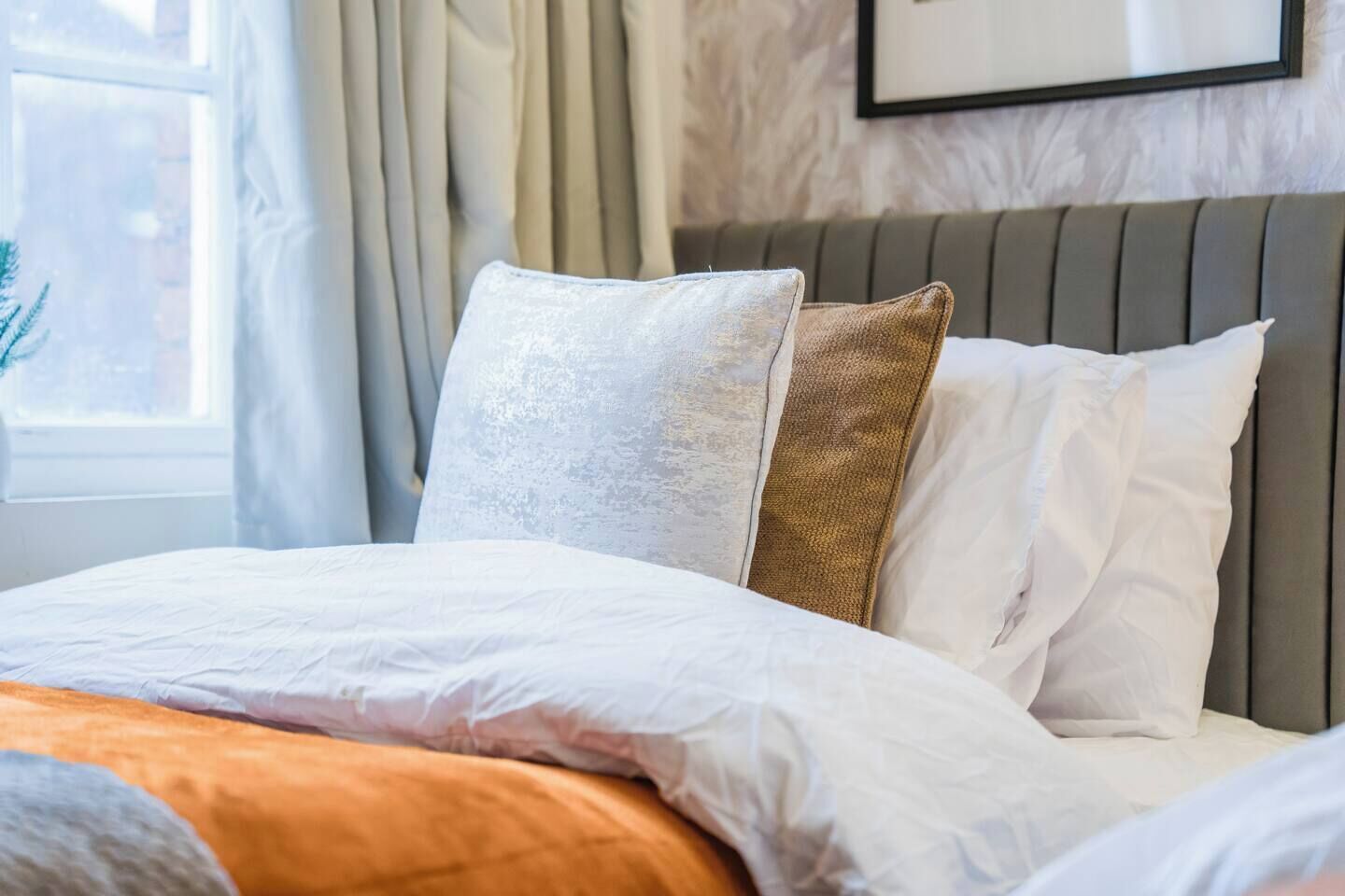 Una cama con almohadas blancas y doradas, un cabecero gris y una ventana con cortinas.