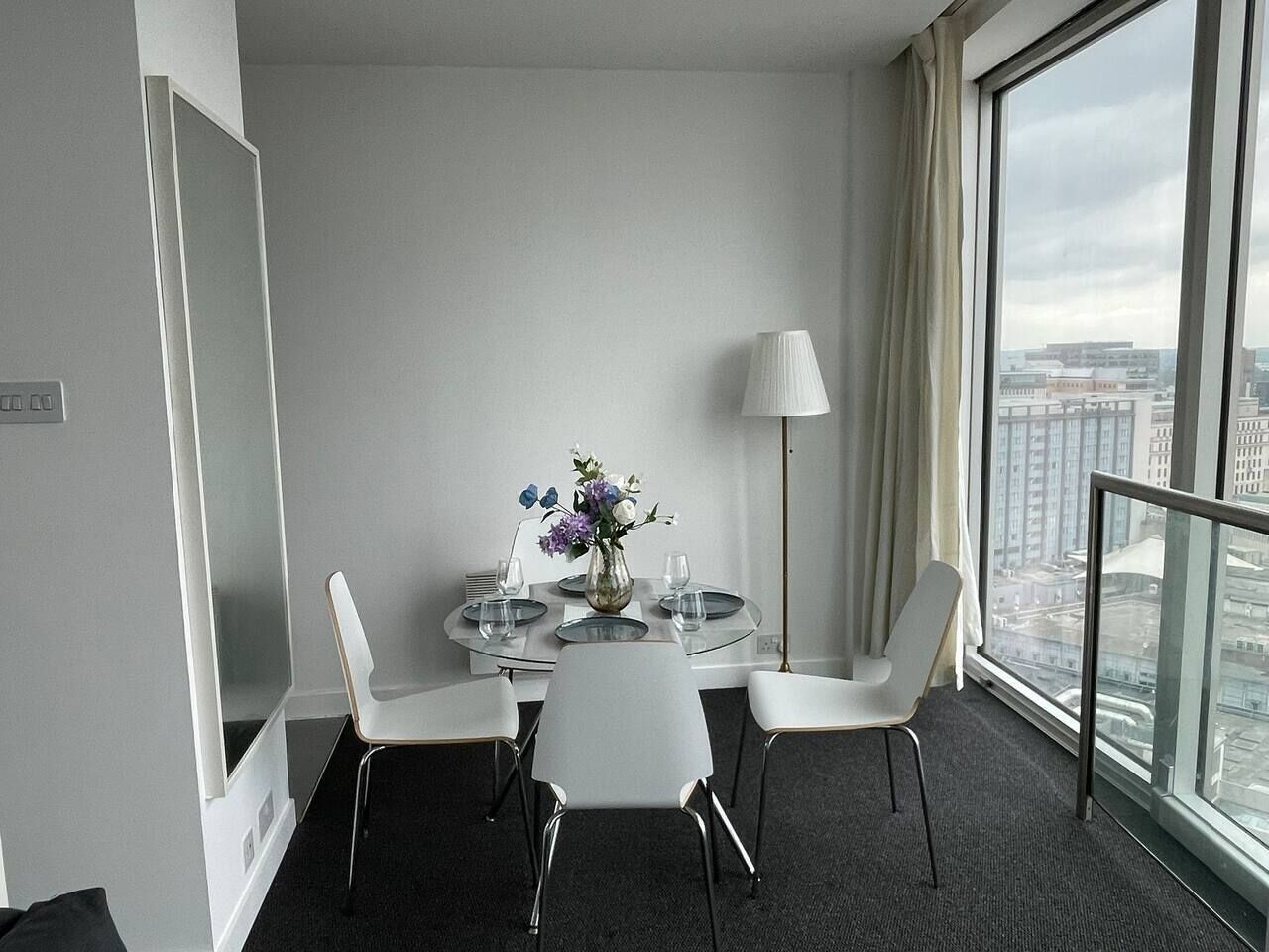 Habitación de hotel con una mesa de comedor redonda, sillas blancas, un jarrón con flores, una lámpara y vistas de la ciudad a través de amplias ventanas.