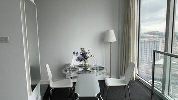 Habitación de hotel con una mesa de comedor redonda, sillas blancas, un jarrón con flores, una lámpara y vistas de la ciudad a través de amplias ventanas.