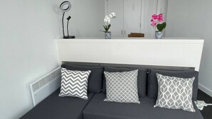 Ein modernes Zimmer mit einem grauen Ecksofa, drei gemusterten Kissen, einem kleinen Tisch mit einer Lampe und zwei Topfpflanzen.