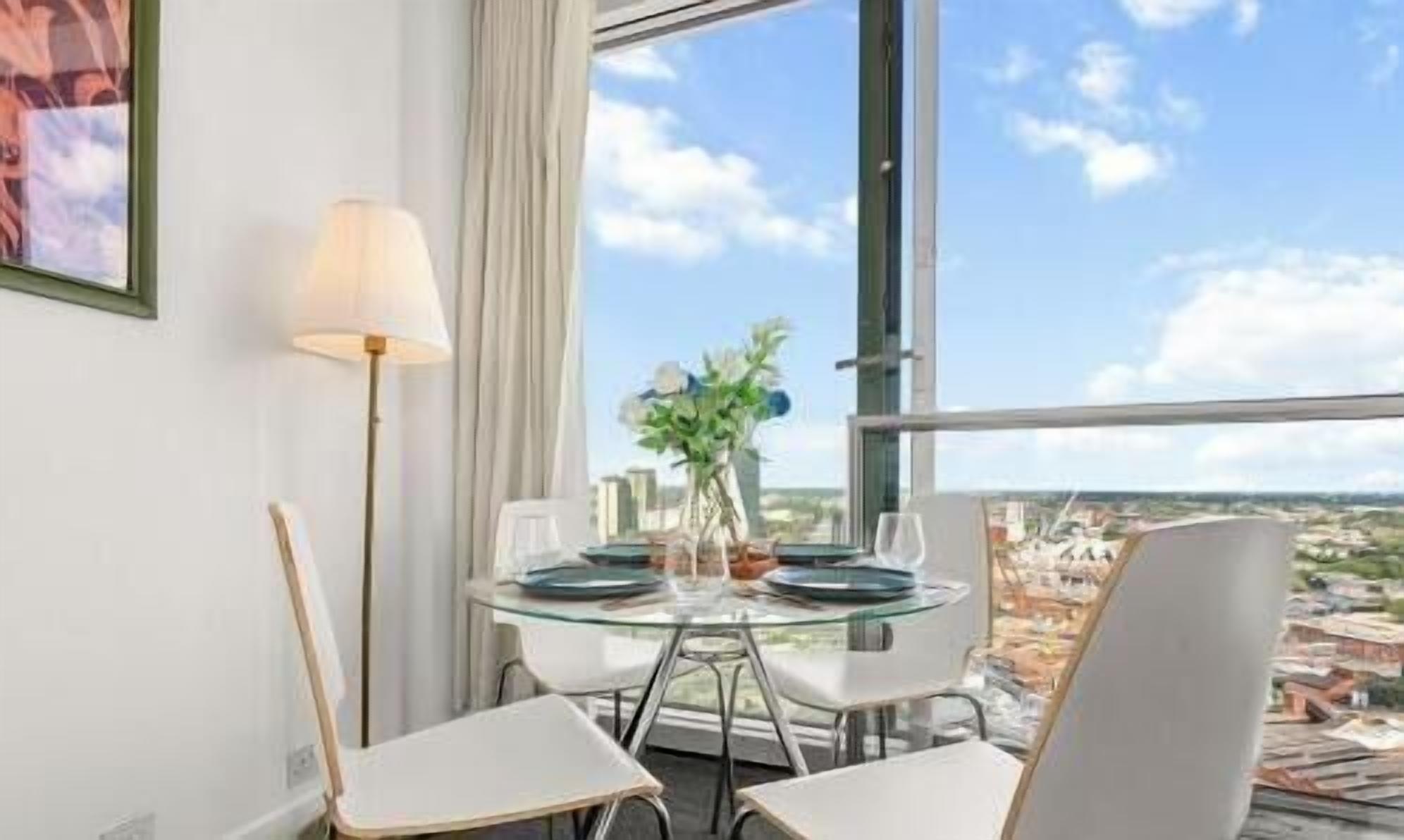 A dining area with a table set for two, a lamp, and a view of the cityscape through large windows.