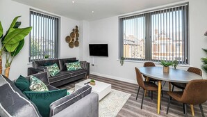 Ein modernes Wohnzimmer mit einer Sofagarnitur, einem Esstisch mit Stühlen, einem Flachbildfernseher und großen Fenstern mit Jalousien.