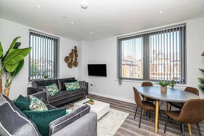 A modern living room with a sofa set, a dining table with chairs, a flat-screen TV, and large windows with blinds. - #95 Crosby Gardens Apartment 2 beds & 1 bath (Waterloo)