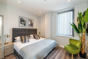 Ein Schlafzimmer mit einem Bett, Nachttischen, einem grünen Sessel, einer großen Pflanze und einem Fenster mit Jalousien.