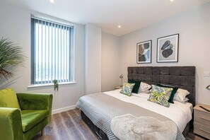 Une chambre à coucher avec un lit, un fauteuil vert, une fenêtre avec des stores et deux tableaux encadrés accrochés au mur.