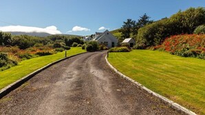 Uma estrada rural que leva a uma casa branca com telhado inclinado, rodeada por vegetação exuberante e um céu limpo.