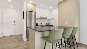 A modern kitchen with a stainless steel refrigerator, a built-in oven, a sink, and a bar area with green upholstered bar stools.