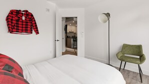 Quarto com uma cama, uma cadeira verde, um casaco vermelho e preto xadrez pendurado na parede, e uma porta que dá para uma cozinha.