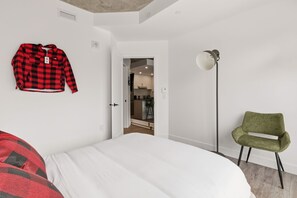 A bedroom with a bed, a green chair, a red and black checkered jacket hanging on the wall, and a door leading to a kitchen. - The Downtown Sugar Shack | Rooftop Pool | Sleeps 8 (Québec)