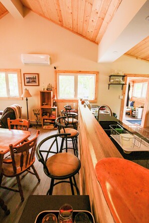 A cozy interior with wooden walls and a high ceiling, featuring a dining area with wooden chairs and tables, a kitchen with a sink and stove, and a window providing natural light.