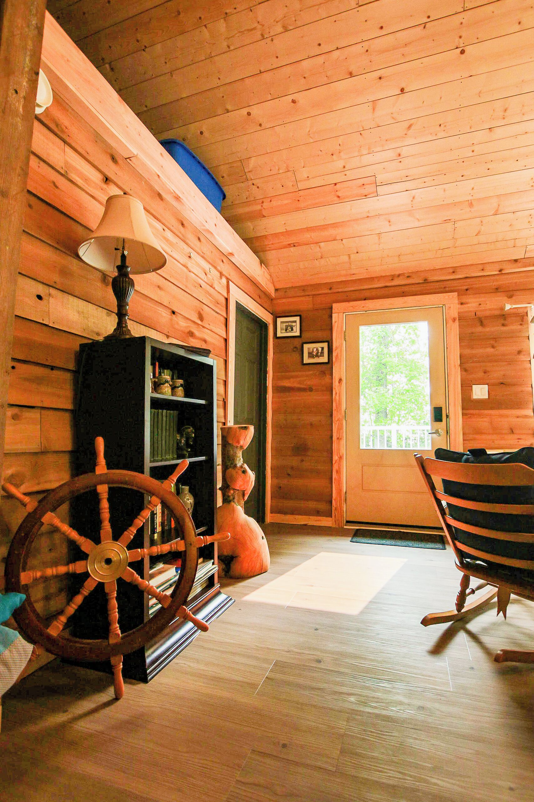 A wooden cabin interior with a rocking chair, a wooden cabinet with books, and a door with a window.