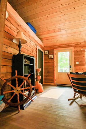 A wooden cabin interior with a rocking chair, a wooden cabinet with books, and a door with a window.