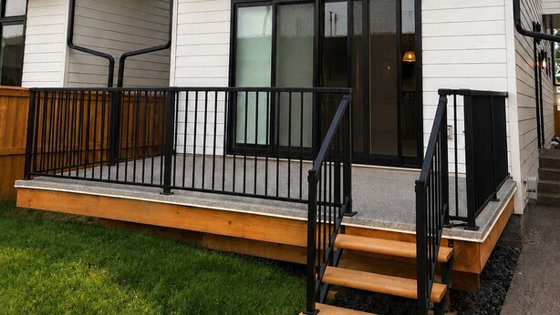 A modern two-story residential building with a flat roof, large windows, and a wooden deck with a black railing.
